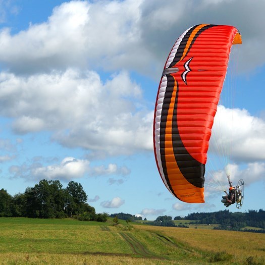 Paramotor pilot flying an orange and red wing over a green field