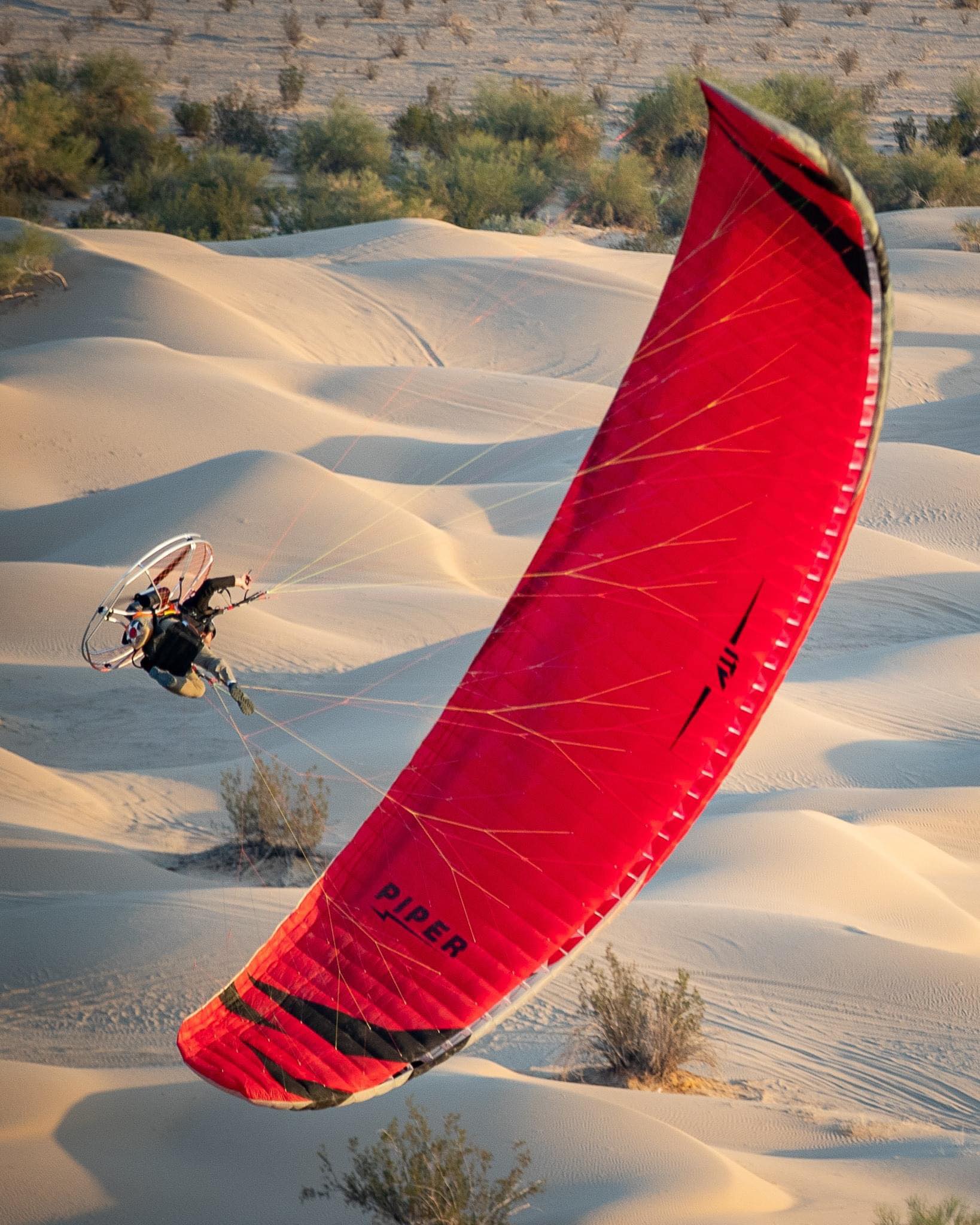 A paramotor pilot flying a red wing low over sand dunes